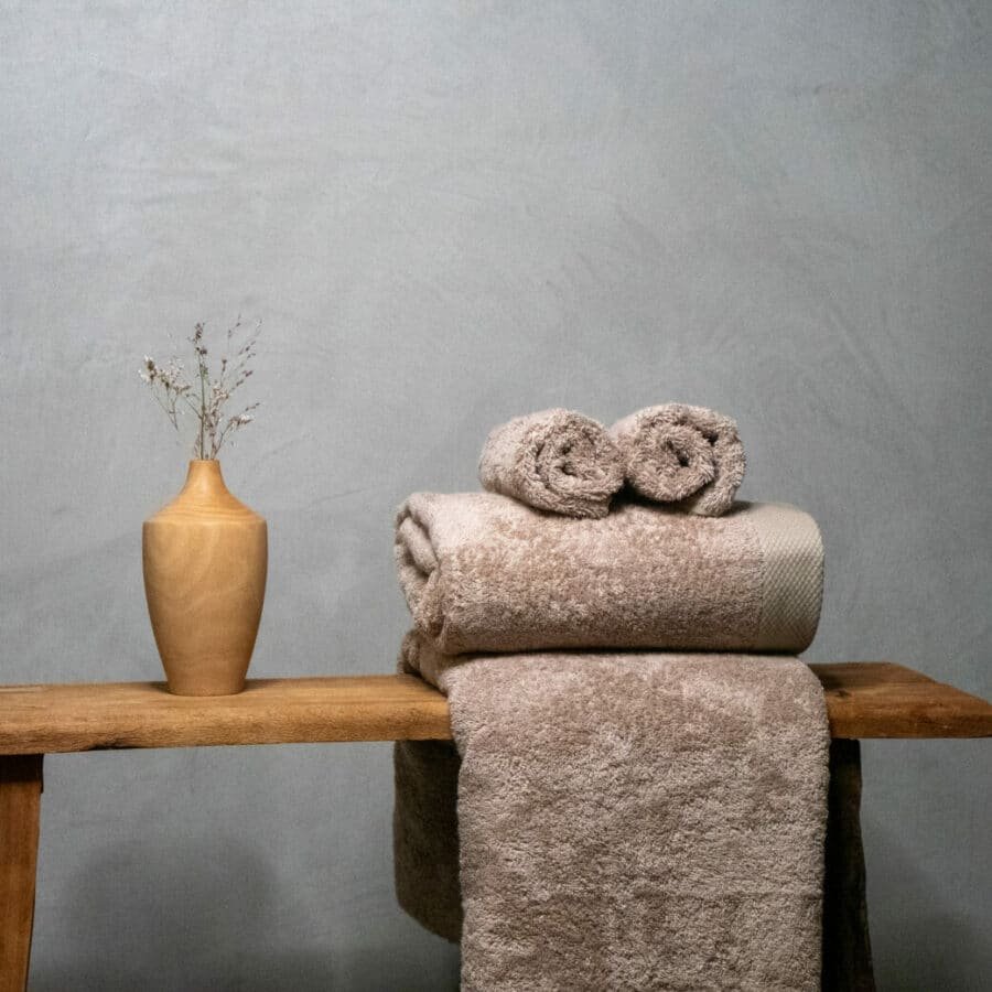 Une serviette en pulpe de bois et fibre de bambou, élégamment disposée sur un banc en bois, ajoute une touche de luxe et de durabilité à cette salle de bain somptueuse. Son aspect naturel et sa texture douce offrent une expérience de confort inégalée, tandis que son engagement écologique ravive l'esprit de bien-être et de respect de l'environnement.